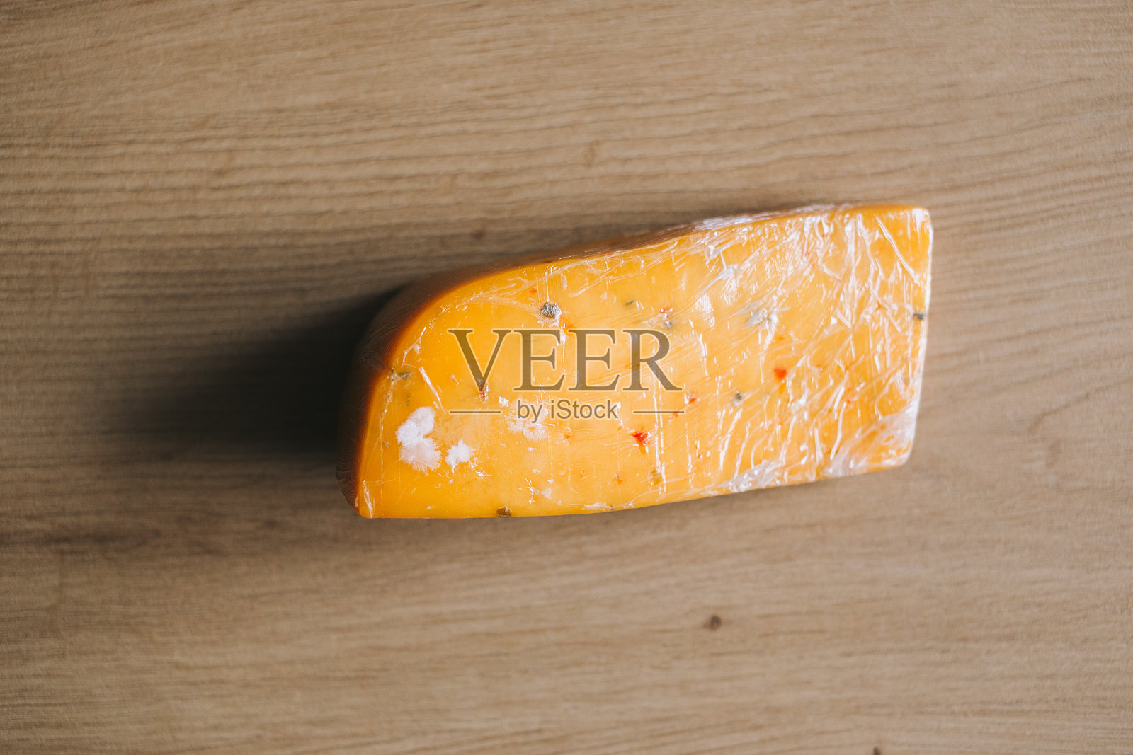 一块用保鲜膜包裹的红色奶酪放在木质表面上。奶酪上有明显的霉点，表明它已经变质。食物储存的缺点。照片摄影图片