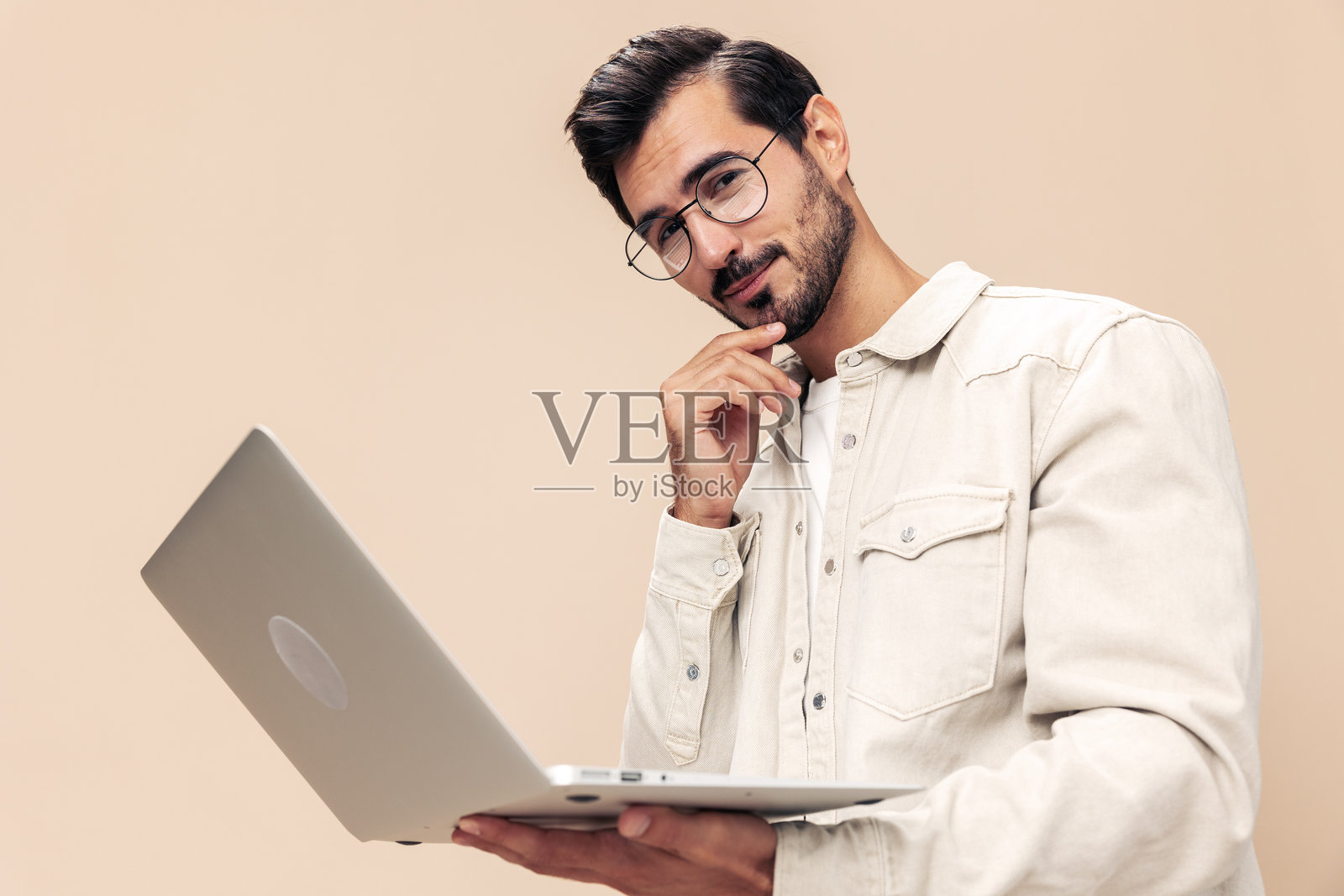 一位时尚男士，戴着眼镜，手持笔记本电脑，神情沉思，穿着白色T恤，背景为米色，展现出时尚的穿衣风格。照片摄影图片