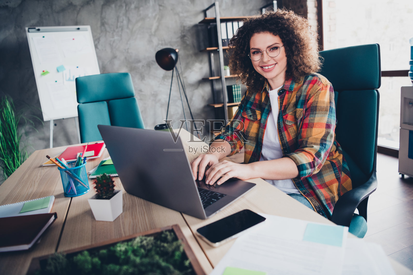 年轻的女商人在现代办公室里工作，使用笔记本电脑，穿着休闲的格子衬衫，卷曲的棕色头发。照片摄影图片