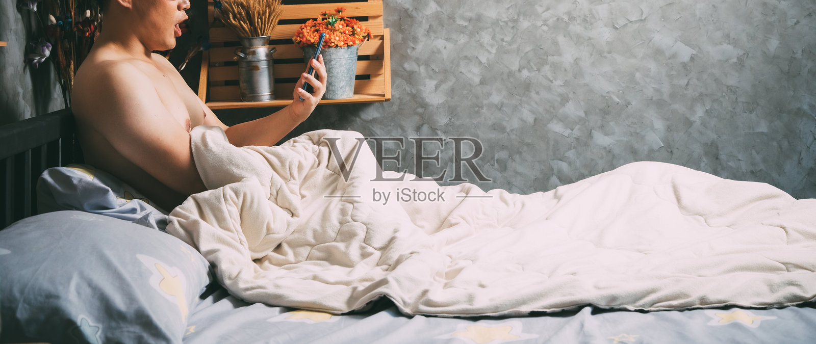 赤裸上身的男子坐在床上，张嘴使用智能手机。照片摄影图片