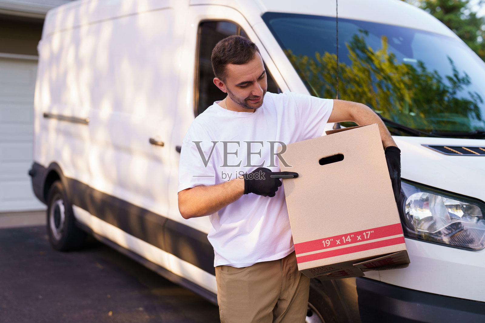 微笑着对着镜头的送货司机，手持并指向一个大箱子，身处大型仓库中。照片摄影图片