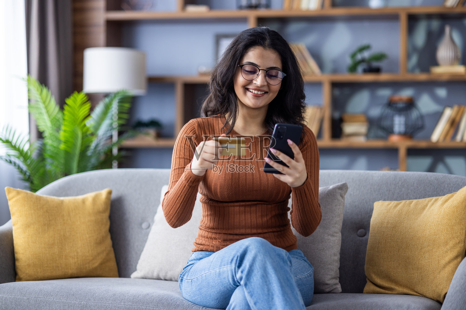 微笑的年轻印度女孩坐在家里的沙发上，使用信用卡和手机。照片摄影图片