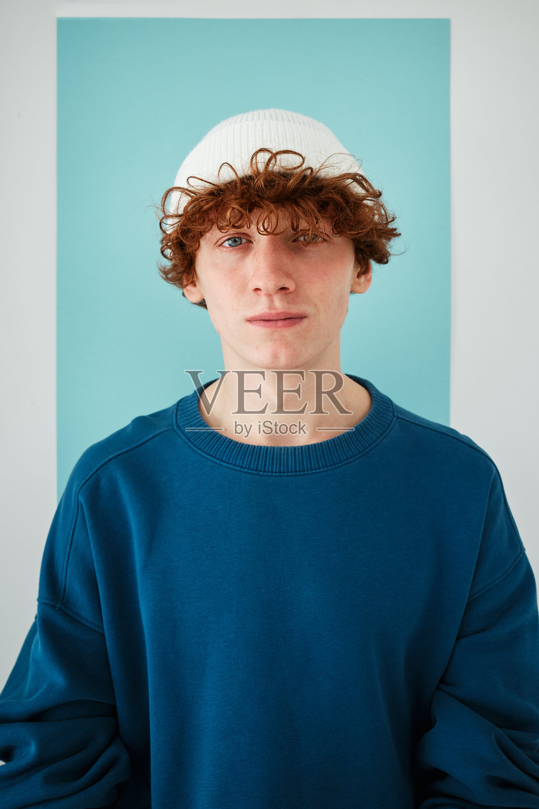 工作室的肖像，一个年轻的红发卷发的家伙在浅蓝色的背景下，全蓝色的衣服照片摄影图片