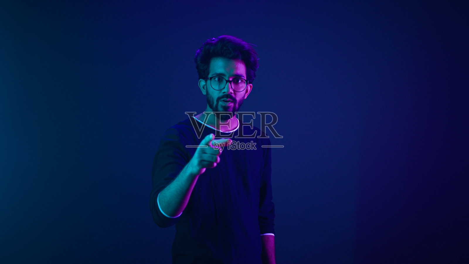 霓虹紫外线背景下，快乐的印度男孩和阿拉伯男工人，程序员黑客计算机工程师，微笑着做出“嘿”的手势，指向镜头，满意地竖起大拇指，支持好主意，表示同意和推荐。照片摄影图片