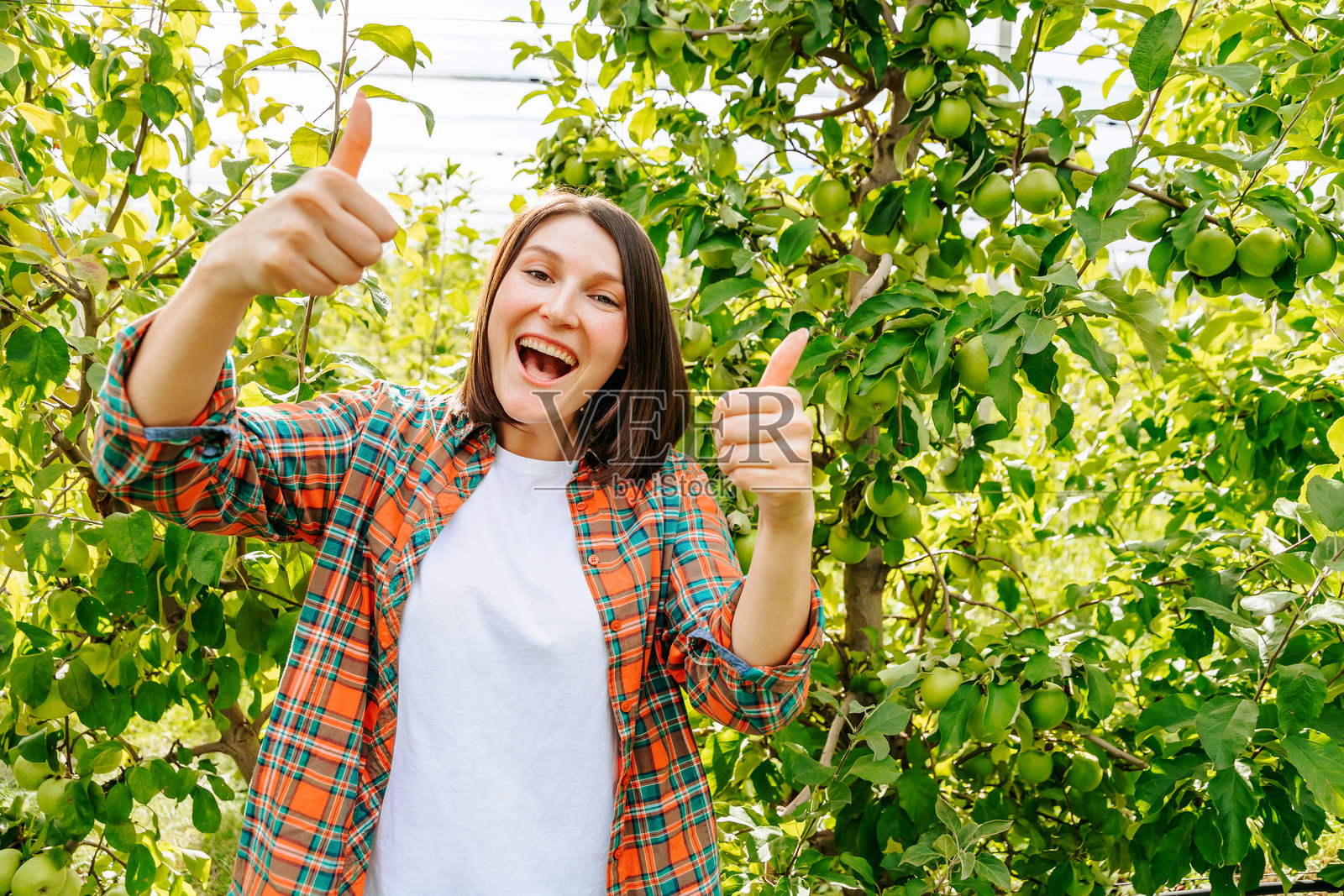 超级情绪化的年轻女子在苹果园里竖起大拇指高兴地喊着什么。照片摄影图片