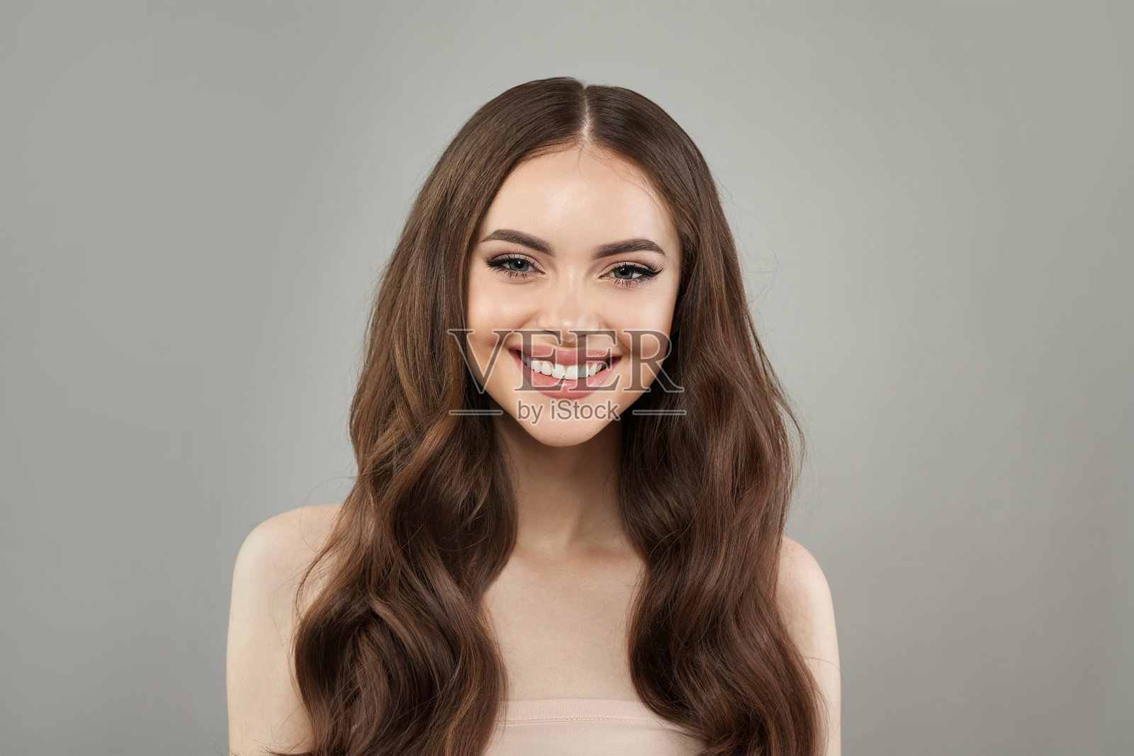 可爱的年轻快乐的女人模特自然化妆，干净健康新鲜的皮肤和长发在灰色背景下摆姿势。卷发，护发，护肤和面部护理的概念照片摄影图片