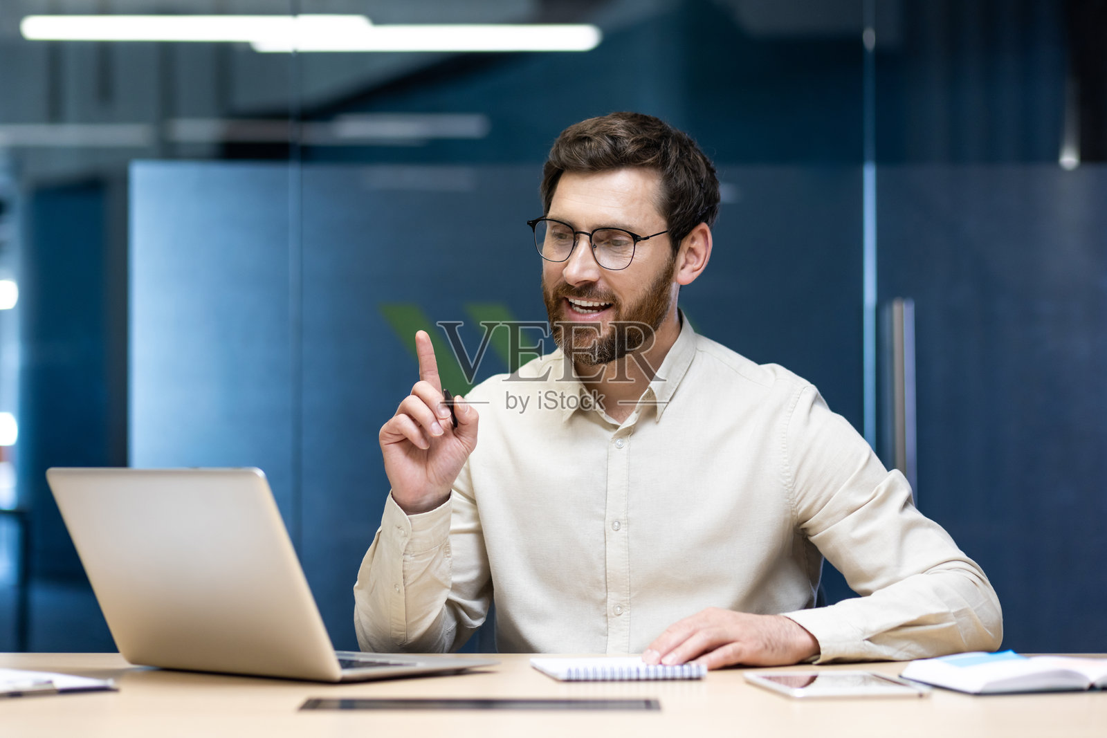 年轻成功的商人在办公室的桌子旁坐着，通过笔记本电脑进行视频通话，手势表达，竖起食指。照片摄影图片