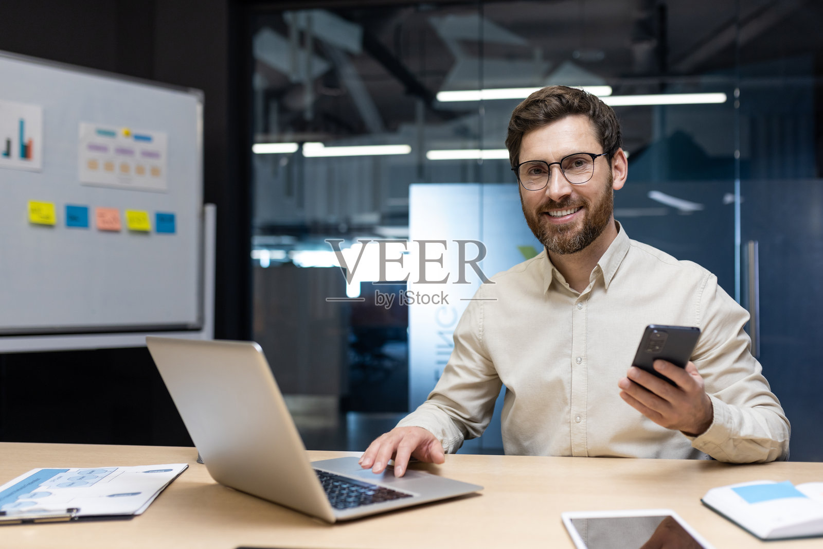 年轻商人在办公室工作，坐在桌子前，手握手机，面带微笑，桌上有一台笔记本电脑和贴满便签的白板。照片摄影图片