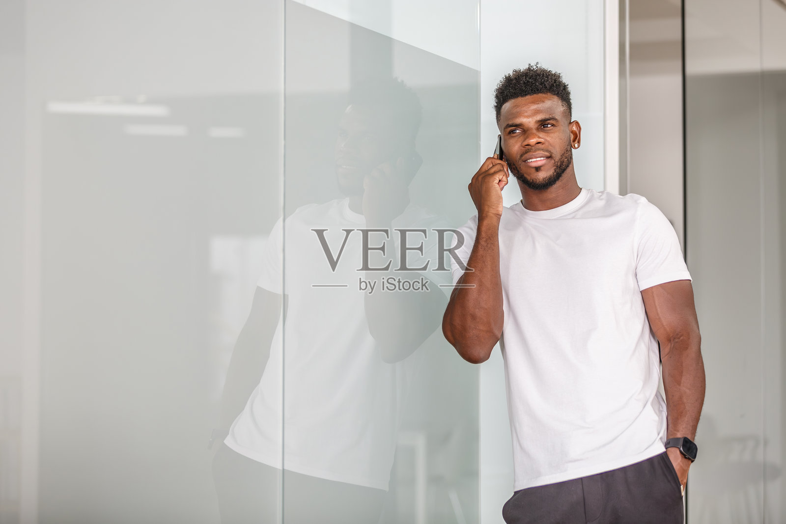 一位年轻男子靠在办公室的玻璃墙上打电话。照片摄影图片