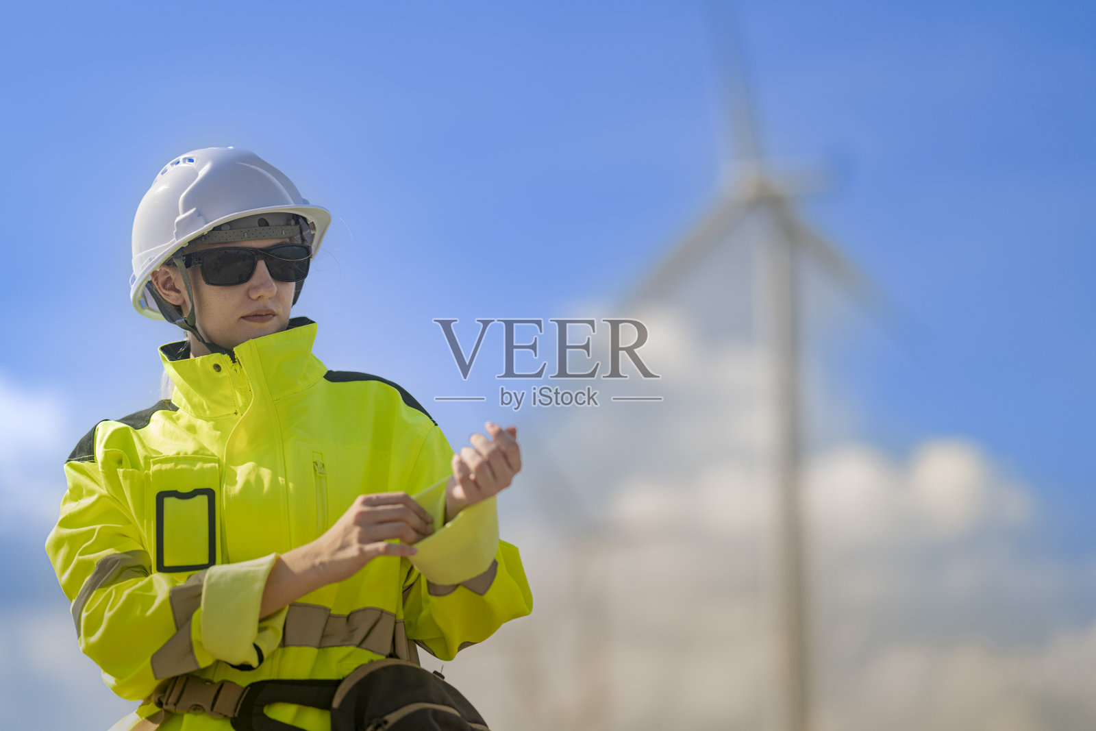 一位穿着高可视性工作服和安全帽的工程师指向背景中的风力涡轮机。这个场景突显了可再生能源以及专业人士在风电场运营中的参与。照片摄影图片