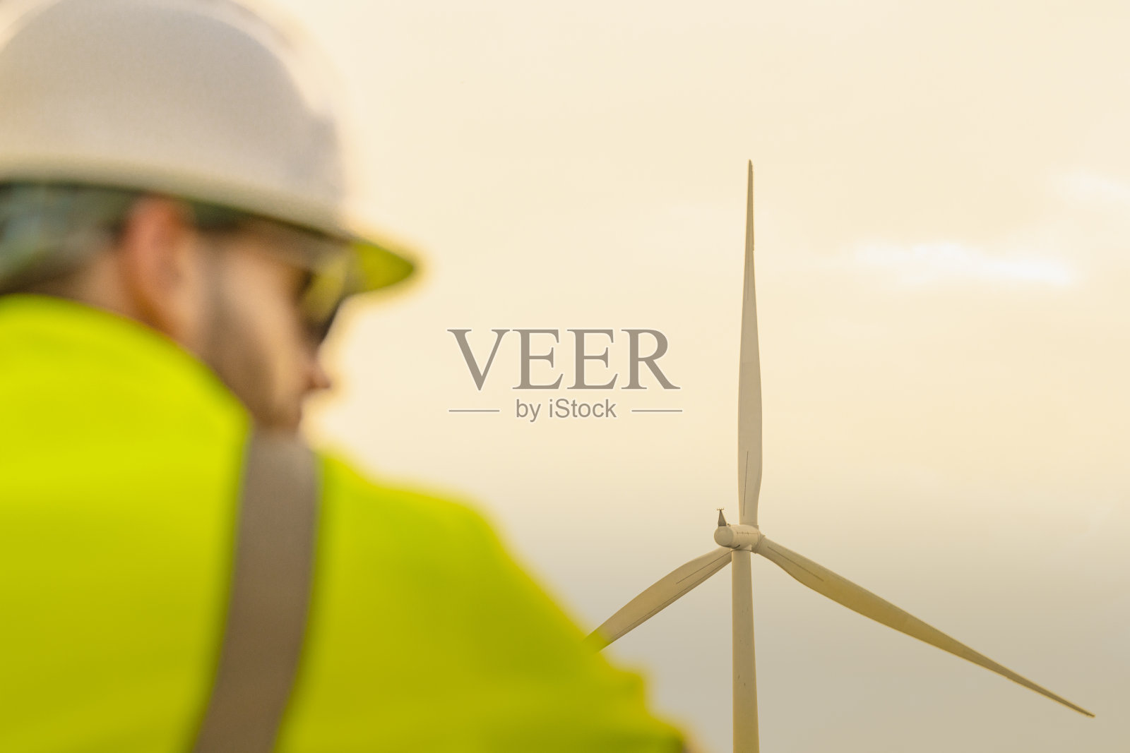 一座风力涡轮机高耸于天空，前景中模糊的工程师穿着高可见度的工作服。该图像强调了可再生能源在维持清洁电力运营中的重要性。照片摄影图片