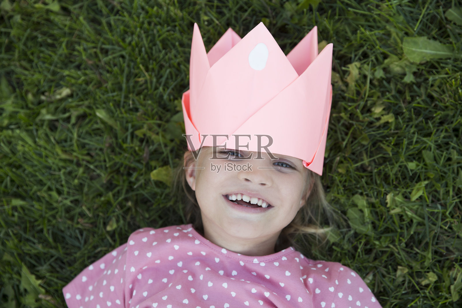 一个带着纸冠笑着的小女孩躺在夏日的绿草地上，看着相机玩得很开心。照片摄影图片