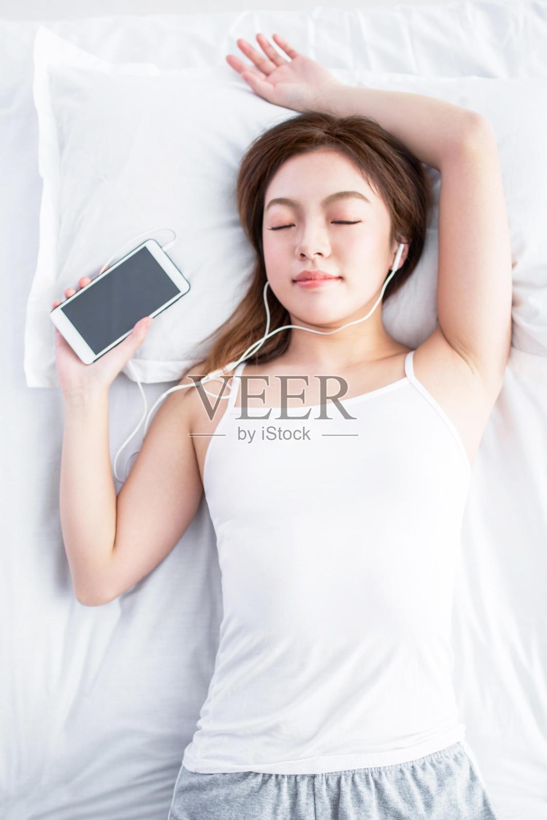 亚洲女性躺在床上，戴着耳机听音乐。照片摄影图片