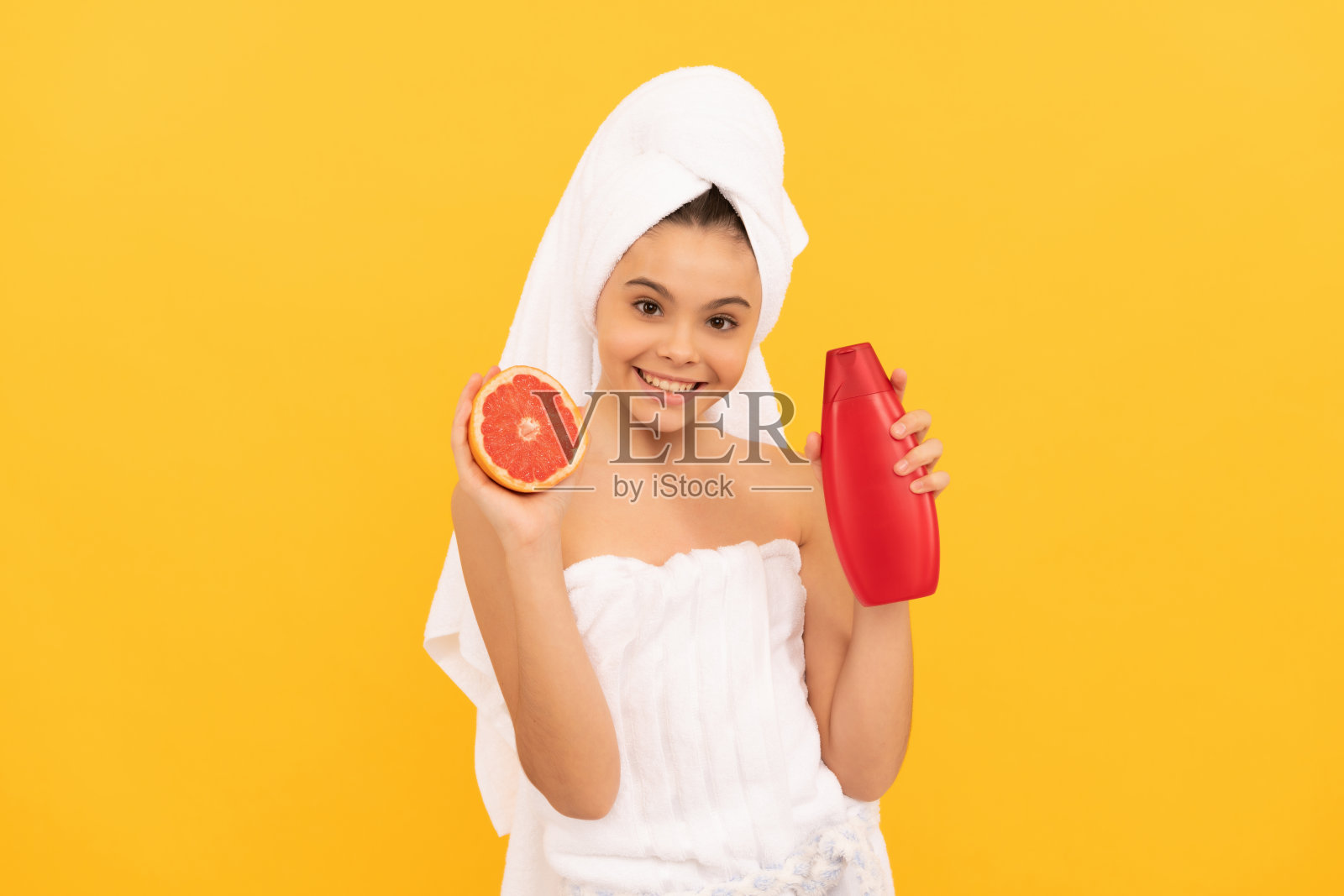 微笑的孩子用毛巾包裹着，旁边有一瓶葡萄柚洗发水，背景为黄色。照片摄影图片
