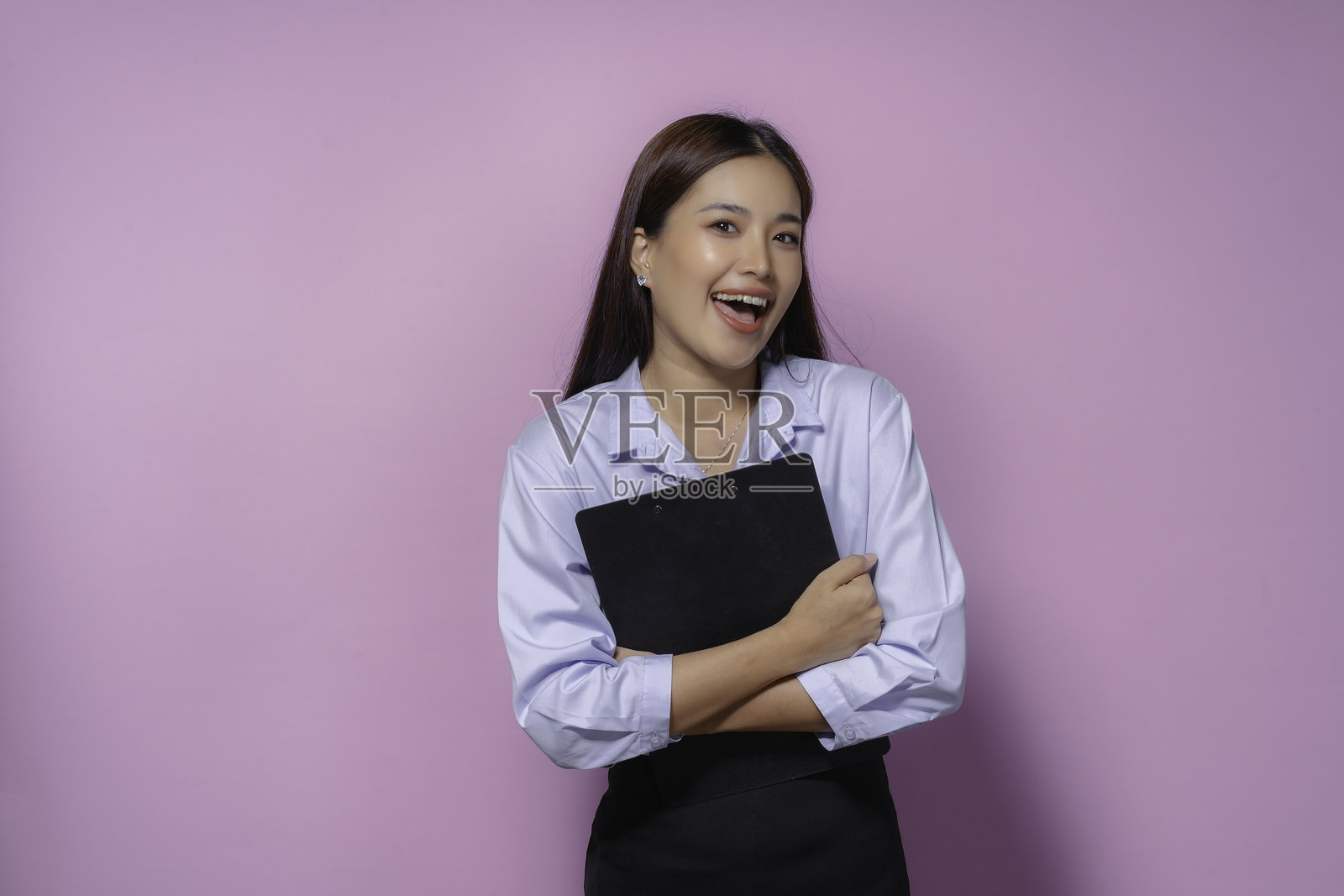 自信的年轻亚洲女商人在工作室拿着写字板微笑着。穿着蓝色衬衫和黑色裙子，头发垂下来。紫色的背景增添色彩，欢快而成功照片摄影图片