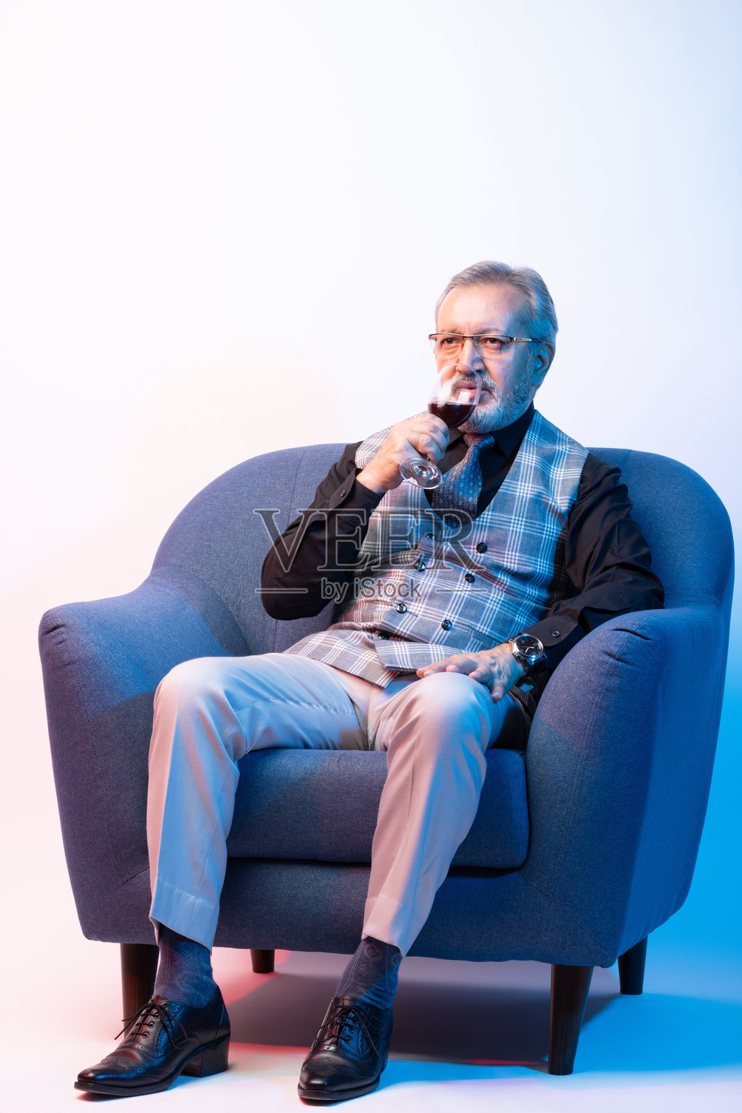 坐在沙发上喝红酒的老年男子，背景为白色。照片摄影图片