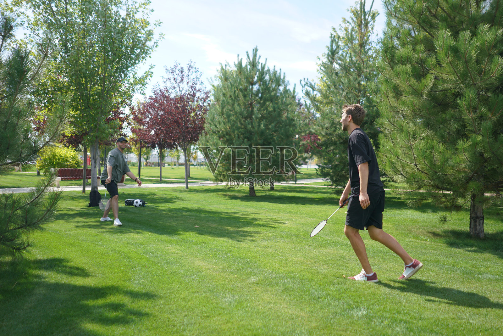 两个人在郁郁葱葱的草坪上进行友好的羽毛球比赛，四周环绕着树木，蓝天晴朗，夏日美好。照片摄影图片