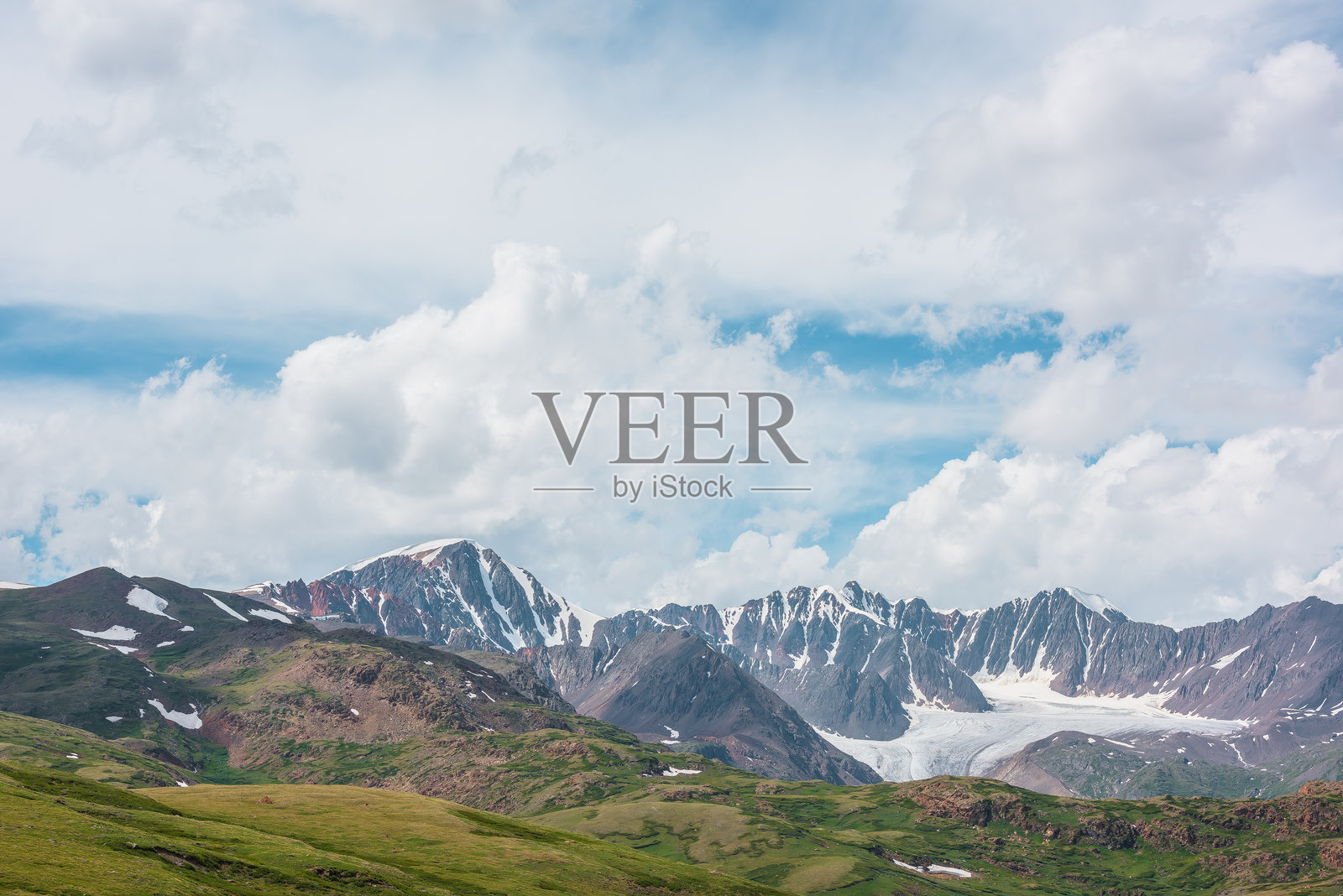 风景如画的阿尔卑斯山景，绿丘与岩石相映，远处可见一座大型雪山峰，周围是红色的尖锐岩石和远处的大冰川，天空中云层变幻，天气多变。照片摄影图片