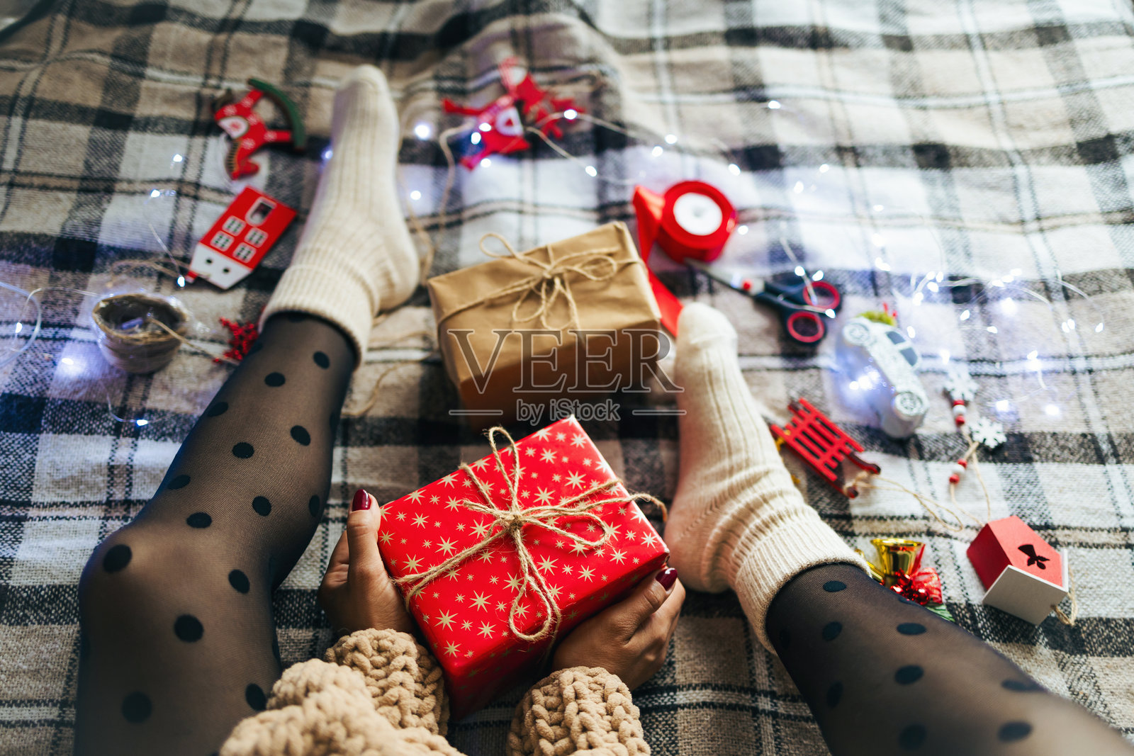 穿着针织毛衣和黑色紧身裤的女人坐在温馨的格子毯子上，旁边有圣诞玩具和包装好的礼盒。圣诞前夕的准备。照片摄影图片
