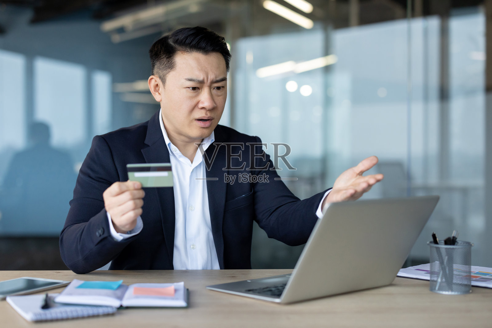 银行账户的财务问题。亚洲年轻男子坐在办公室的桌子旁，手握信用卡，失望地看着笔记本电脑，双手摊开。照片摄影图片