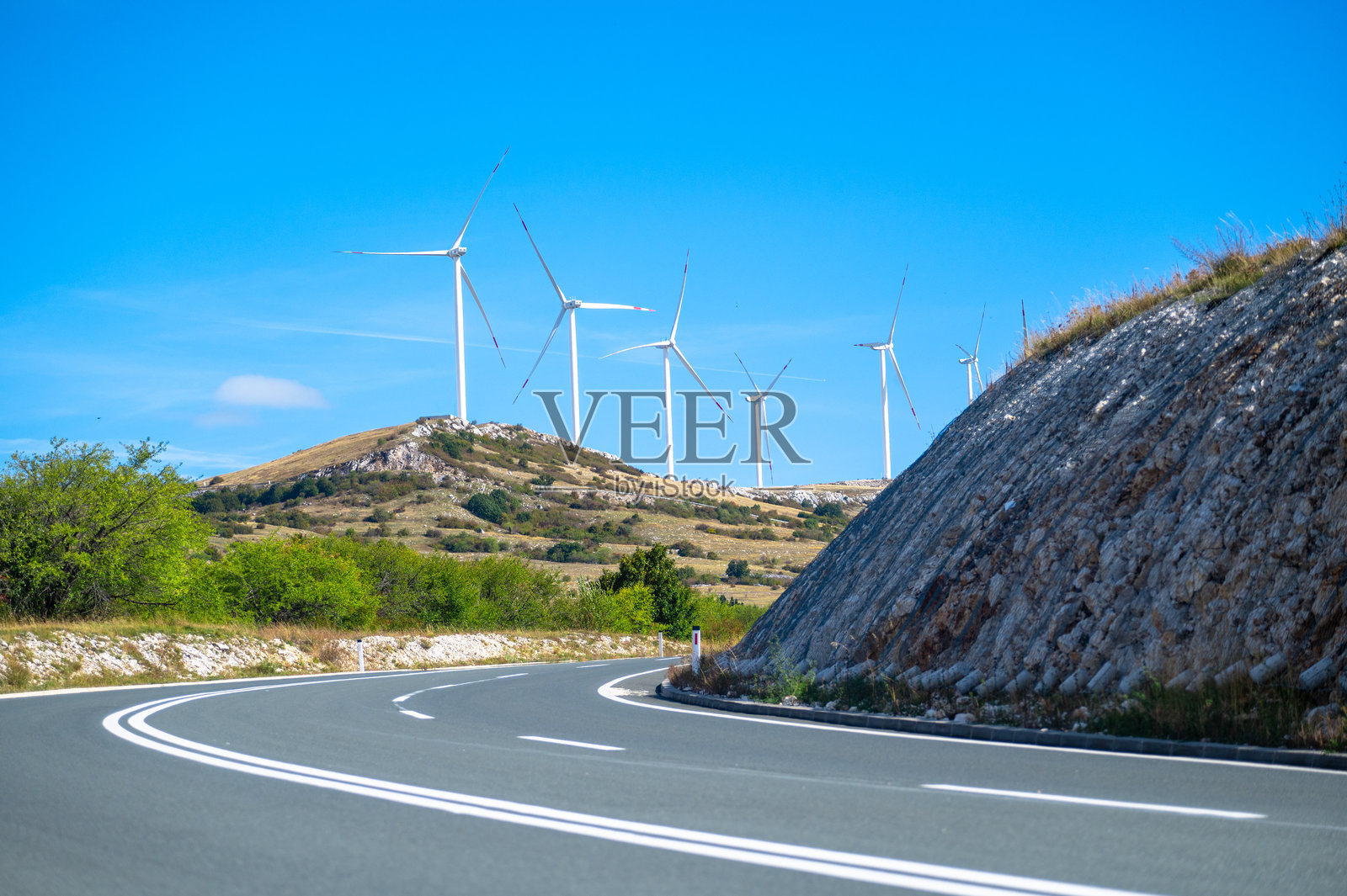 一条道路通向一座山丘，山丘上有多个现代风力涡轮机，指引着通往地平线上一个大型风电场的方向。照片摄影图片