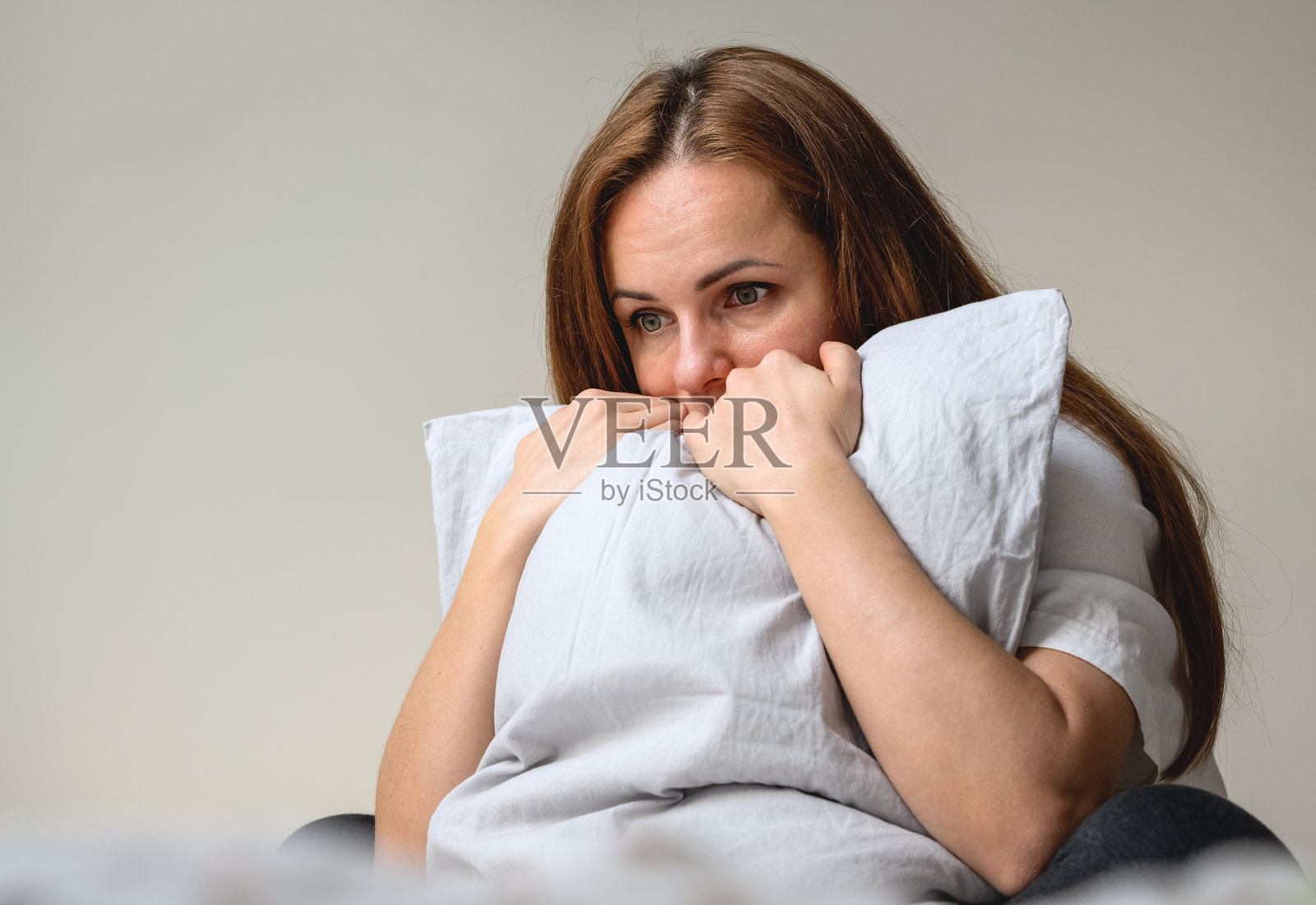 不快乐的女人坐在卧室的床上，抱着枕头。照片摄影图片