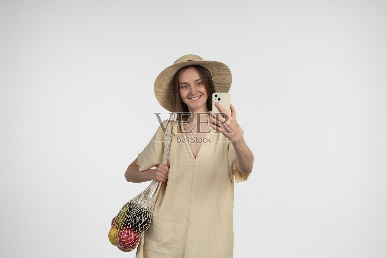 一位穿着轻便服装和太阳帽的女性在明亮的室内空间中微笑着自拍，手里拿着一个装满五彩水果的可重复使用的购物袋。照片摄影图片