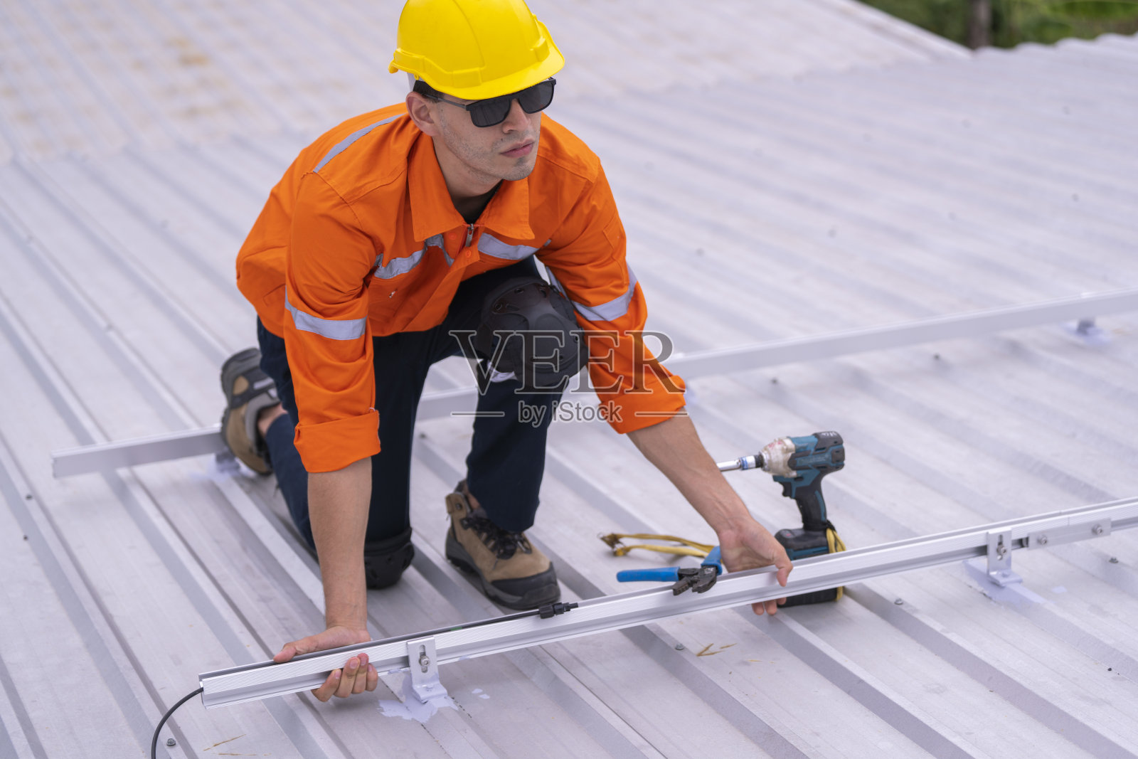 穿着安全制服和头盔的技术人员跪在屋顶上，使用电钻进行精确的安装工作。这个场景突显了建筑行业中的安全性、专业性和技术专长。照片摄影图片