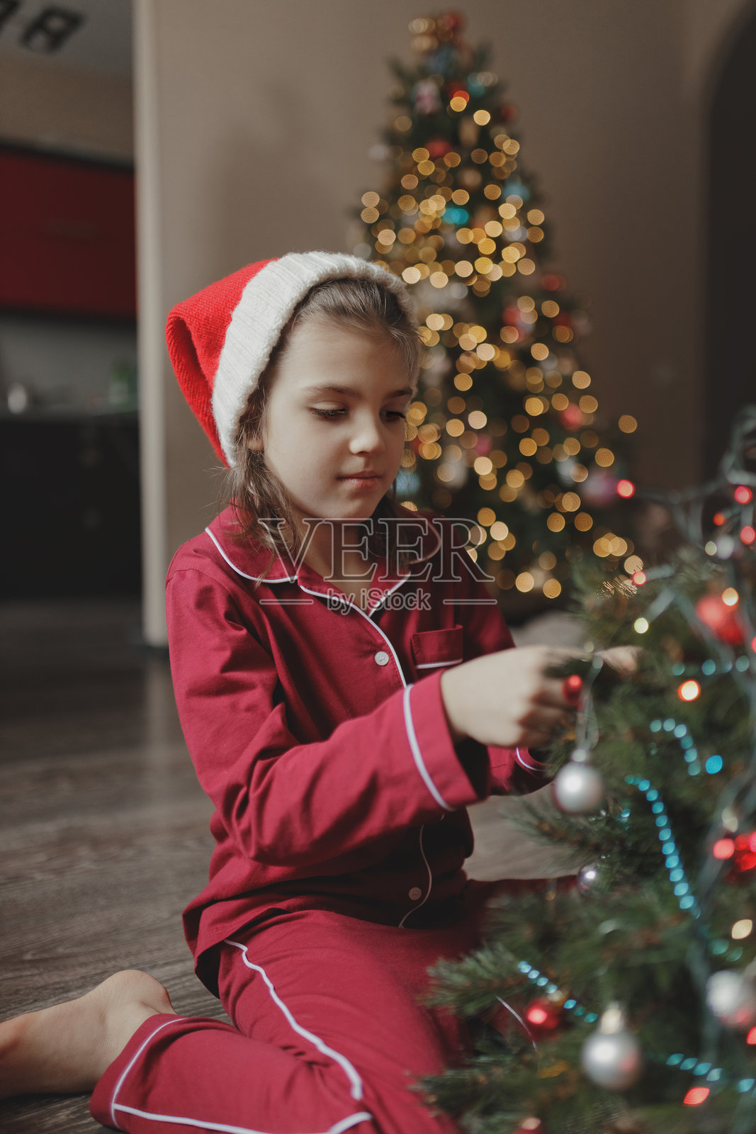7岁女孩穿着红色睡衣和圣诞老人帽，坐在客厅地板上装饰圣诞树。照片摄影图片