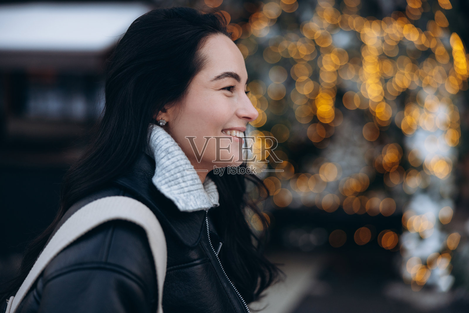 年轻微笑女性在城市街道上，靠近装饰有闪亮灯串的圣诞树。照片摄影图片