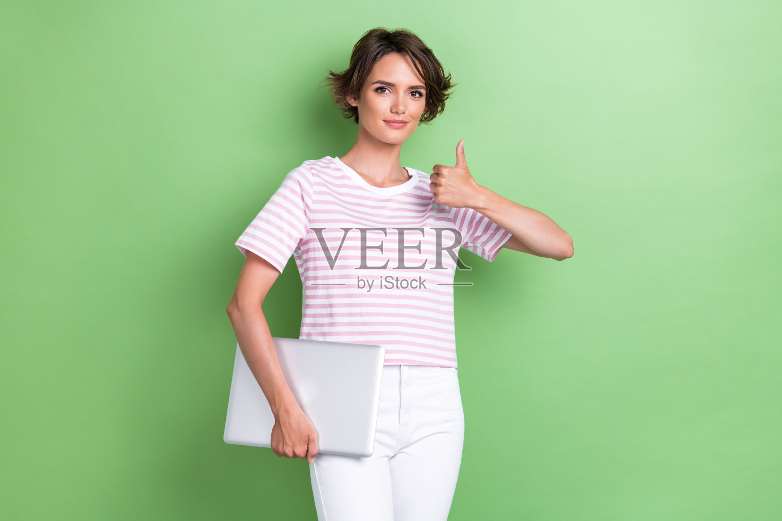 年轻迷人、可爱、自信的女孩穿着粉色T恤，竖起大拇指，手持笔记本电脑，展现出良好的品牌表现，背景为绿色。照片摄影图片