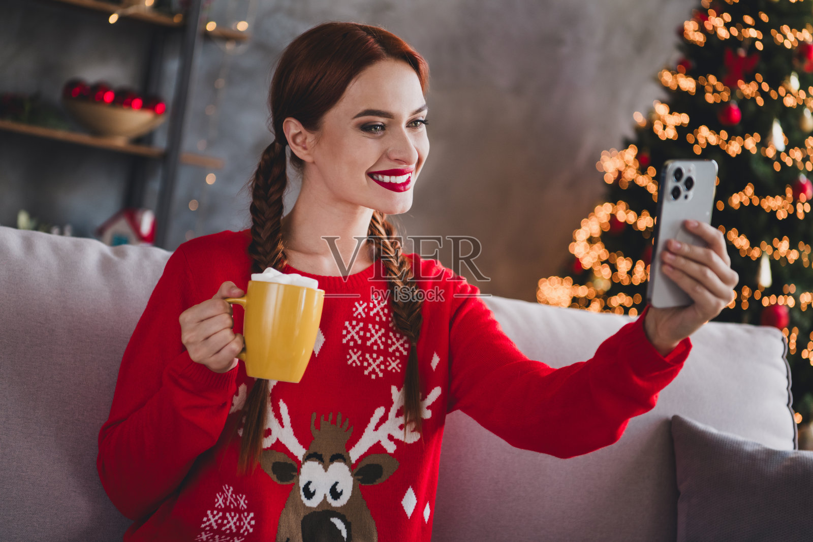 可爱的年轻红发女性在家中派对装饰的室内环境中，穿着圣诞服装，喝着可可，自拍照。照片摄影图片