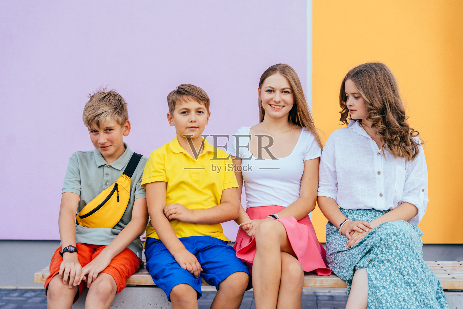 四个孩子朋友坐在长椅上，背景是五彩斑斓的墙壁。照片摄影图片