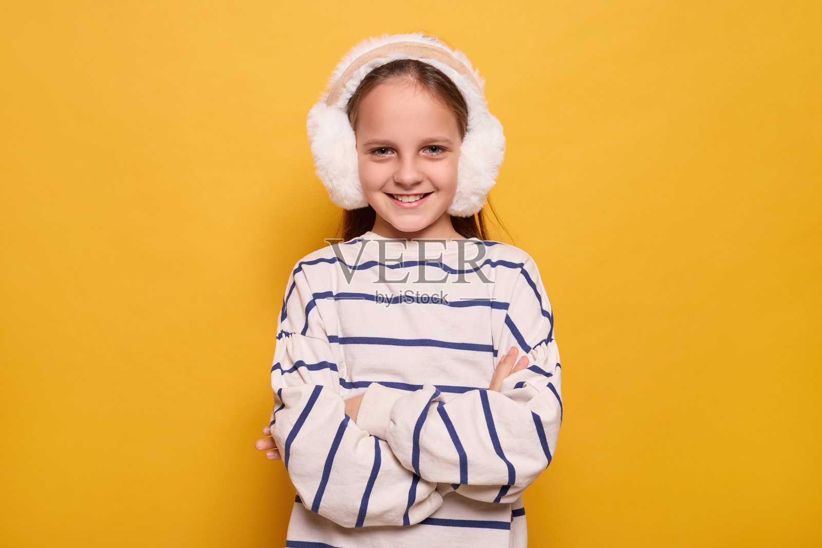 开心的小女孩戴着毛绒耳机，自信地微笑，双臂交叉，展现出活泼友好的精神，散发出温暖的快乐，背景为黄色。照片摄影图片