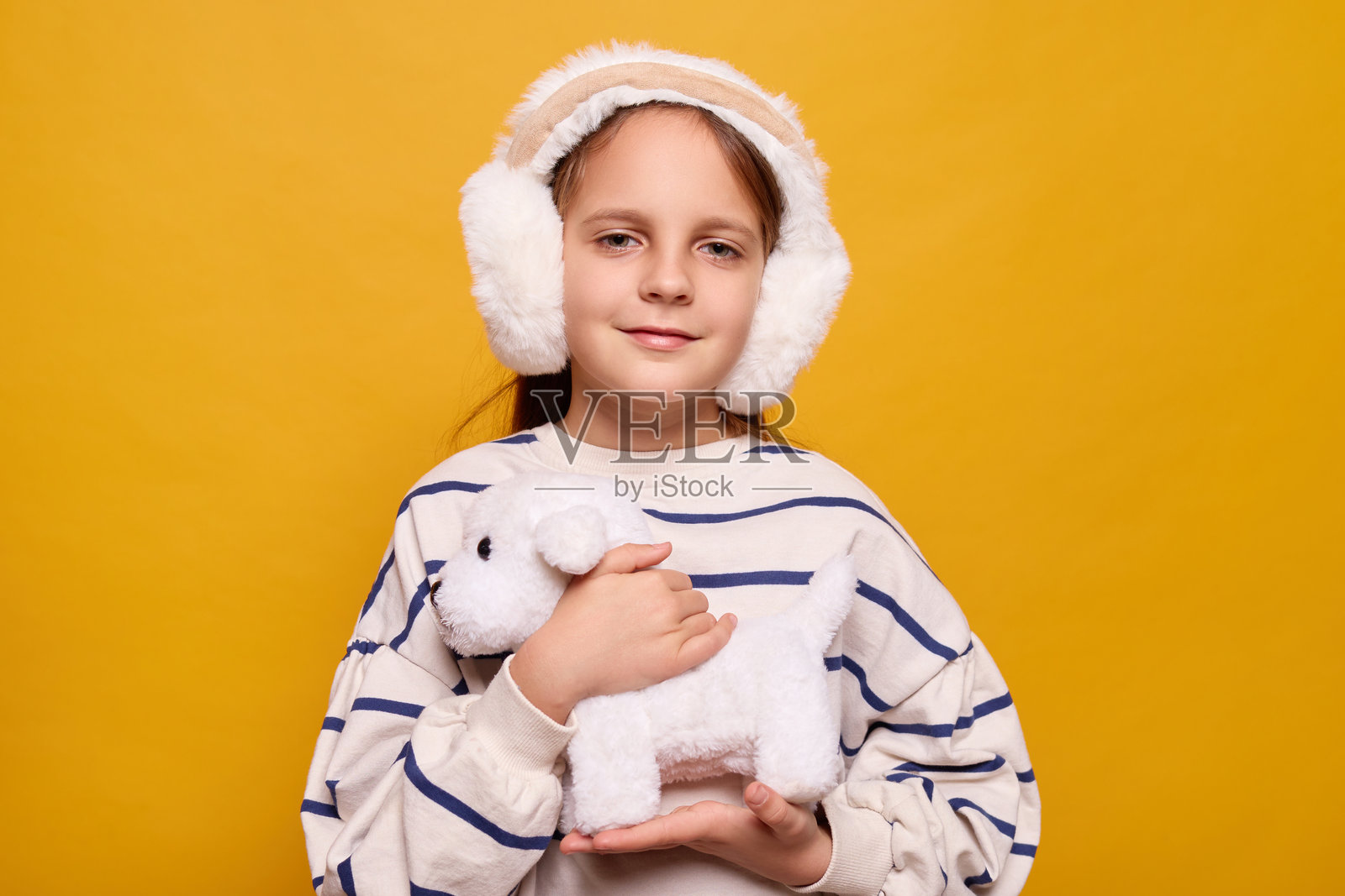 开心的小女孩穿着条纹衬衫，戴着毛绒耳机，微笑着接过一只柔软的白色小狗作为礼物，满足地拥抱着它，在室内与玩具一起玩耍，背景为黄色。照片摄影图片