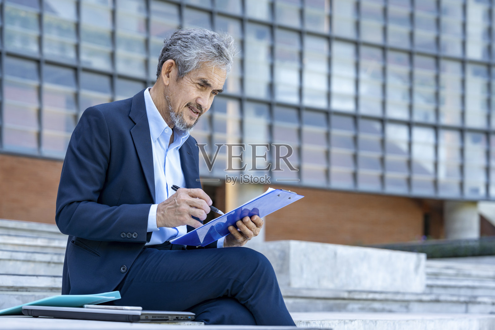 穿着西装的老练商人在户外建筑旁休息时随意工作，坐着写文件。照片摄影图片