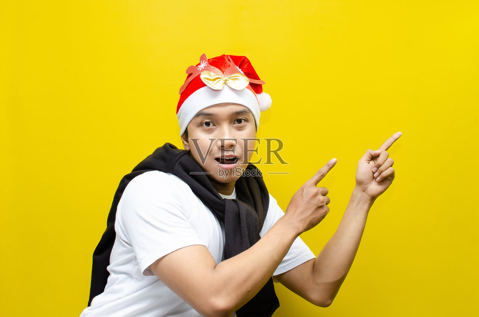 快乐的亚洲男子侧面图，戴着圣诞老人帽，手指指向空白处，双手展示着某物。照片摄影图片