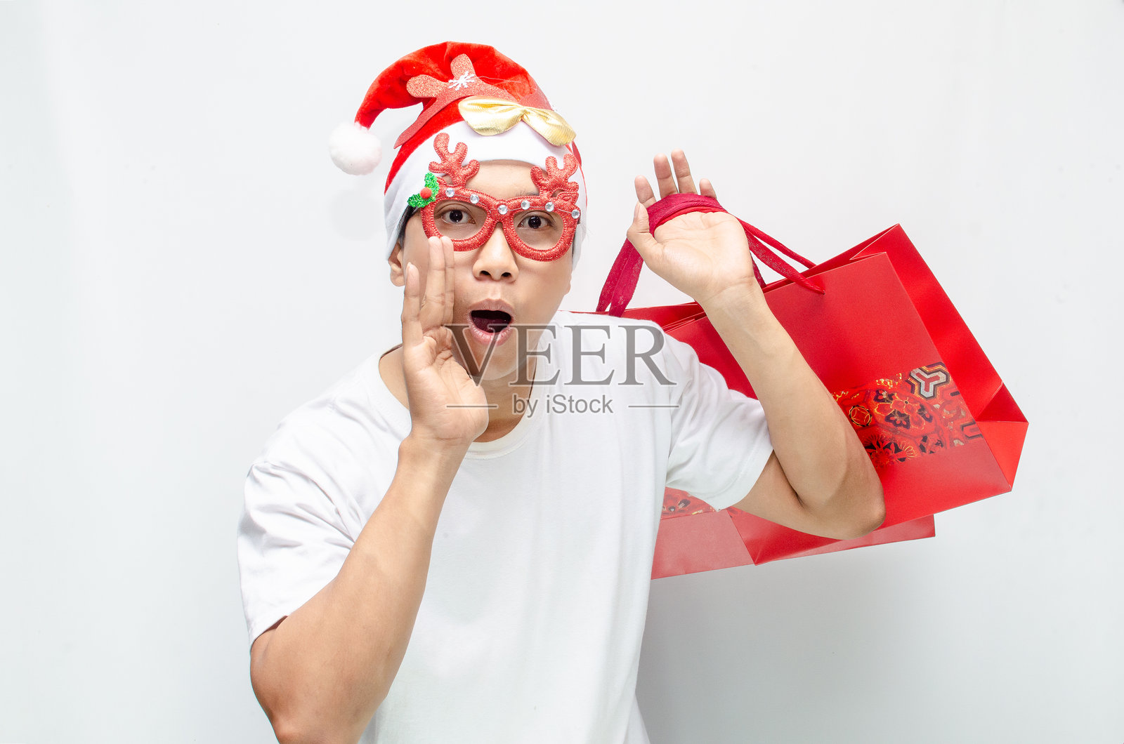 兴奋的年轻亚洲男子戴着圣诞老人帽和圣诞眼镜，手提红色购物袋。照片摄影图片