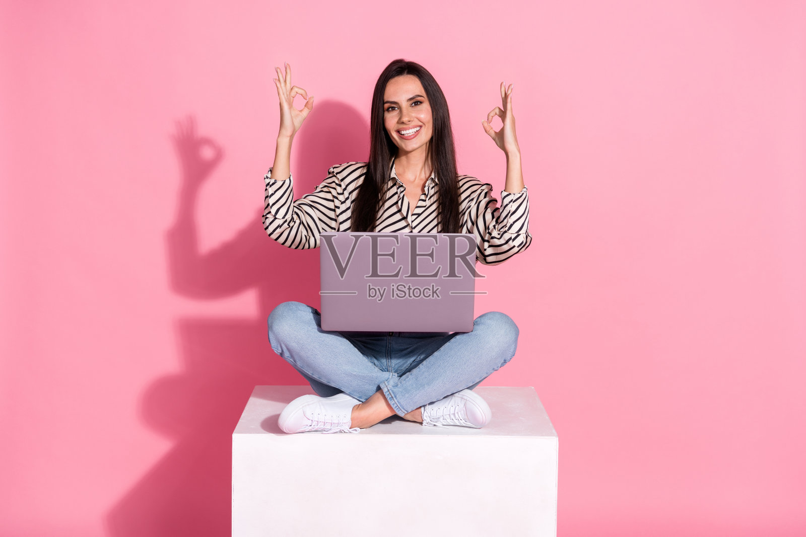 年轻女性坐在白色方块上，背景为粉色，手持笔记本电脑，做出“OK”手势，适合广告或购物主题。照片摄影图片