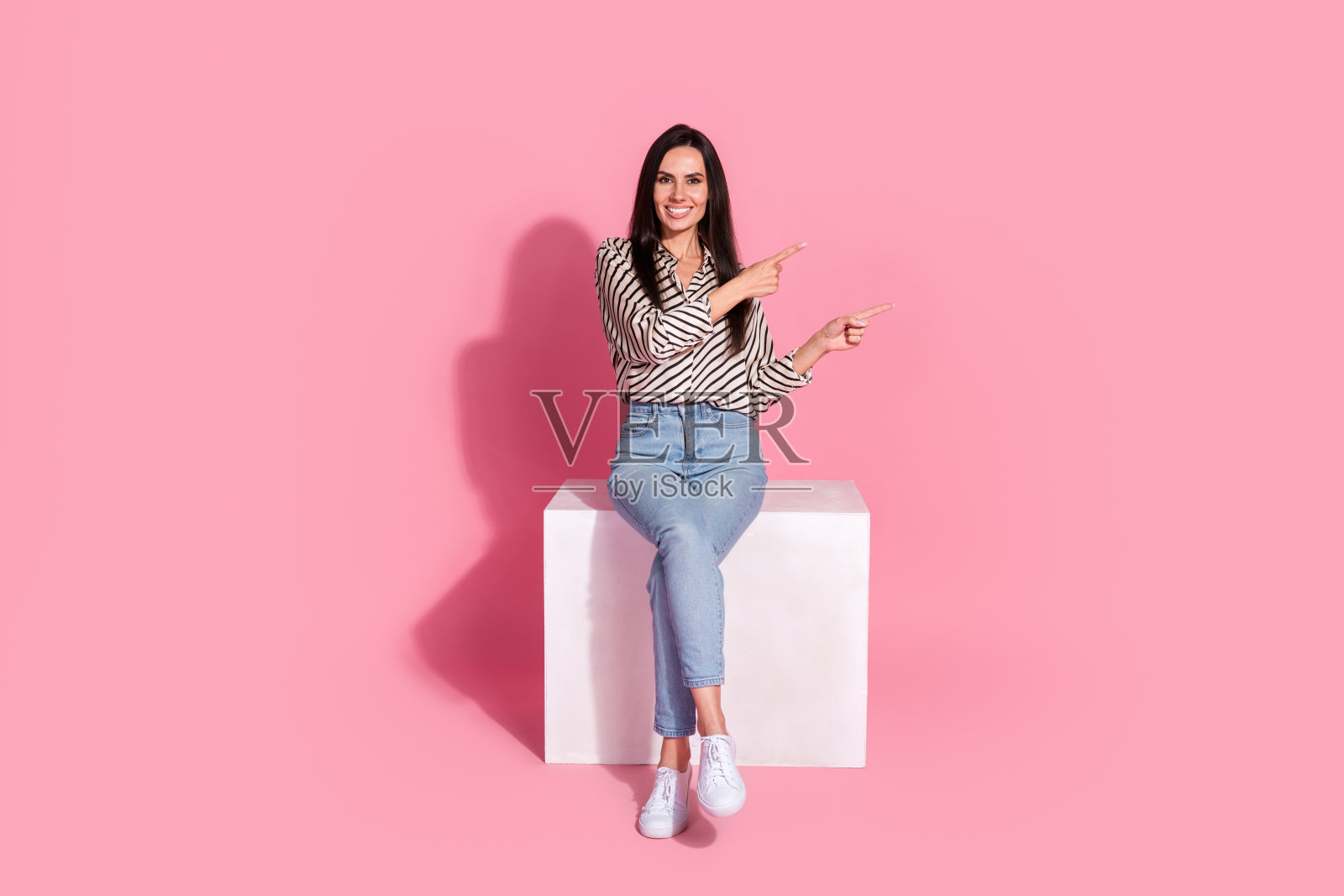 自信的年轻女性穿着条纹衬衫，坐在白色立方体上，背景为粉色，面带愉快的表情，指向一侧，非常适合广告和宣传材料。照片摄影图片
