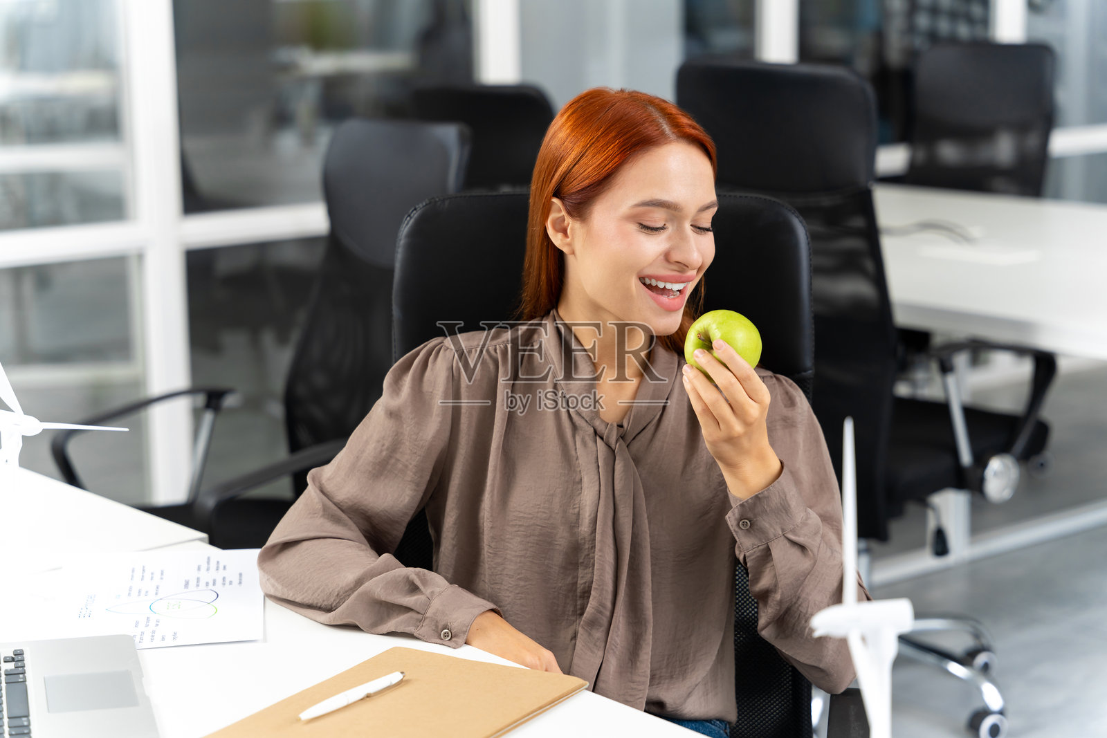 微笑的红发年轻女性手握苹果坐在办公室里照片摄影图片