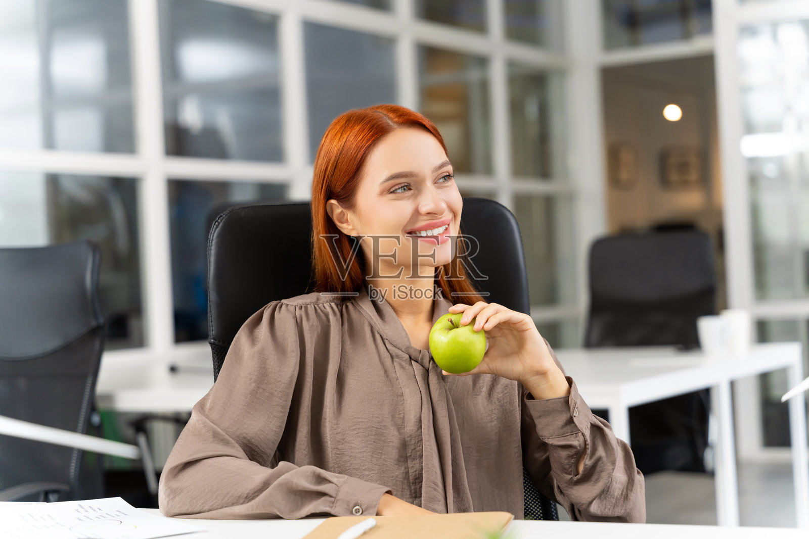 漂亮的微笑红发年轻女性手握苹果坐在办公室里照片摄影图片