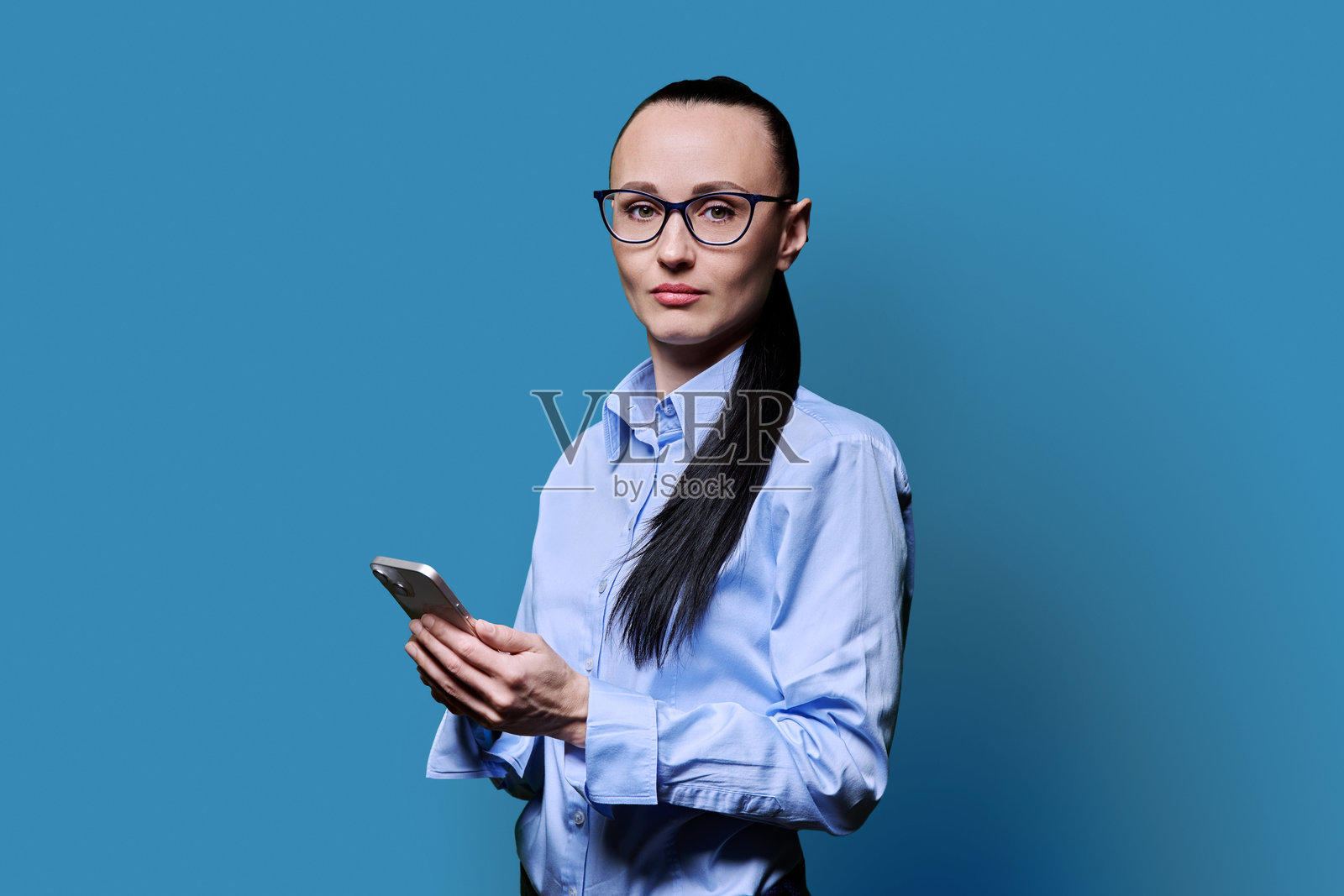蓝色背景下，商务女性手持智能手机对着镜头微笑。照片摄影图片