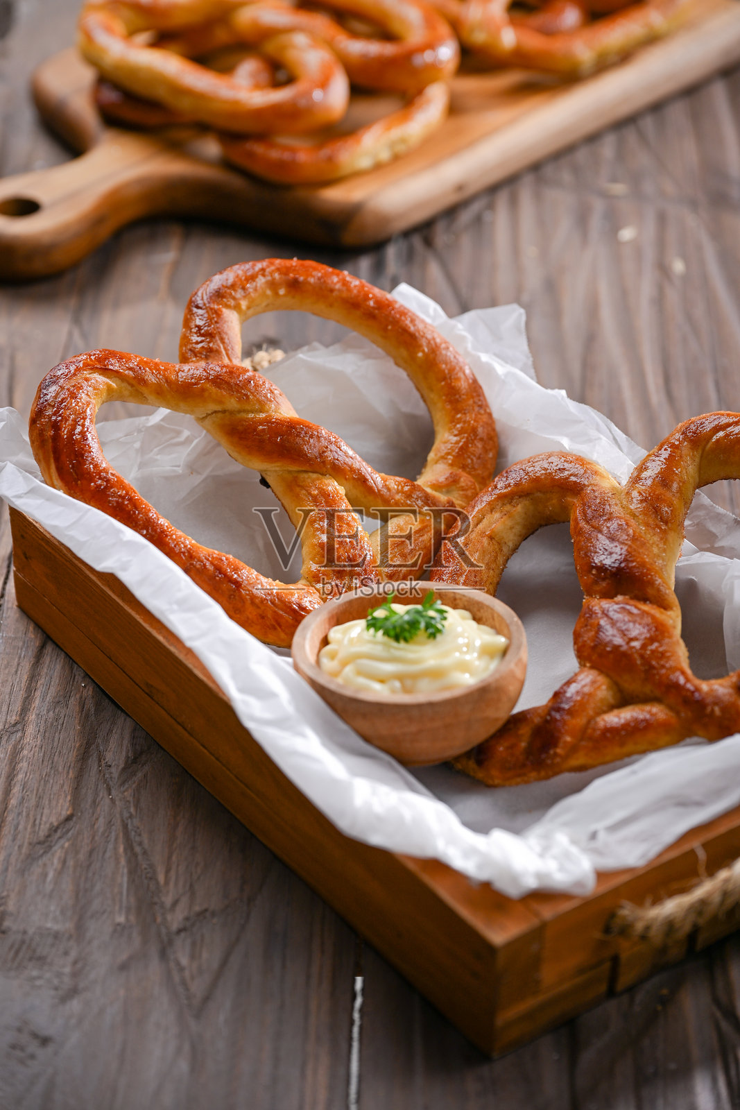 一个温暖的金棕色椒盐卷饼旁边放着一小碗奶油蘸酱，上面点缀着香菜。粗犷的木质背景和羊皮纸让这个小吃散发出自制的艺术气息。照片摄影图片