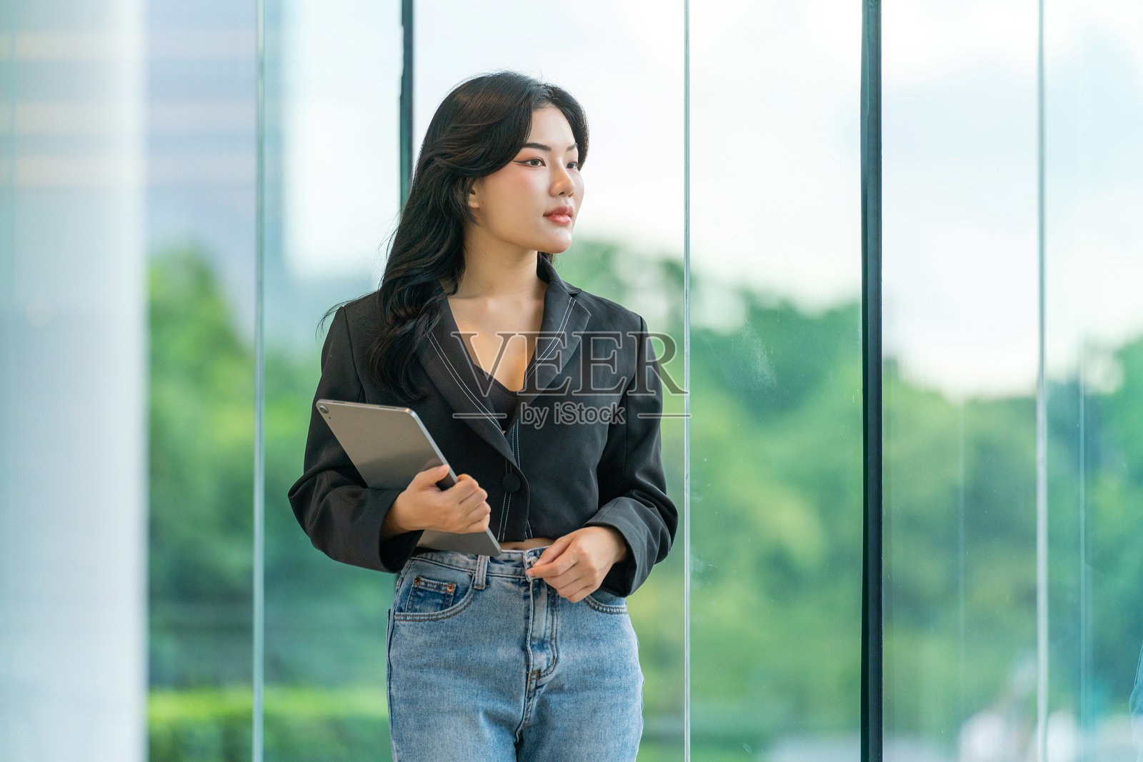 穿着黑色西装的亚洲女性在大型玻璃窗前手持数字平板，窗外绿意盎然，神情满足地望向一旁。照片摄影图片