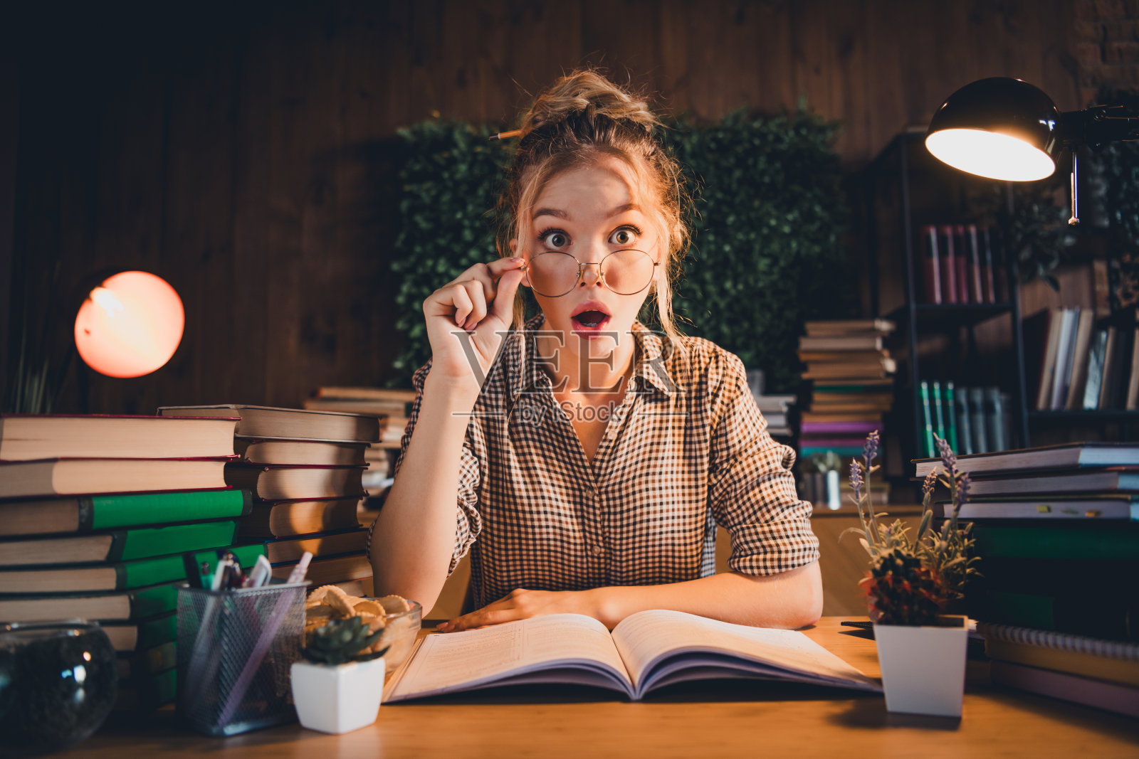 年轻女子穿着格子衬衫，惊讶地在家中深夜学习，周围堆满了书籍。照片摄影图片