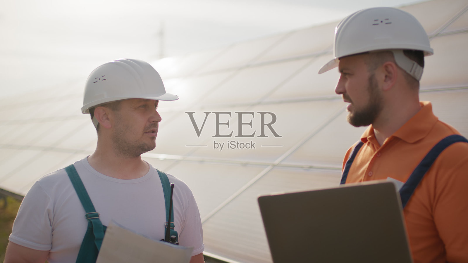 不同种族的男性工程师在太阳能农场讨论高效的施工计划。两名穿着防护头盔和制服的男性正在交谈并查看平板电脑屏幕。太阳能公园。照片摄影图片
