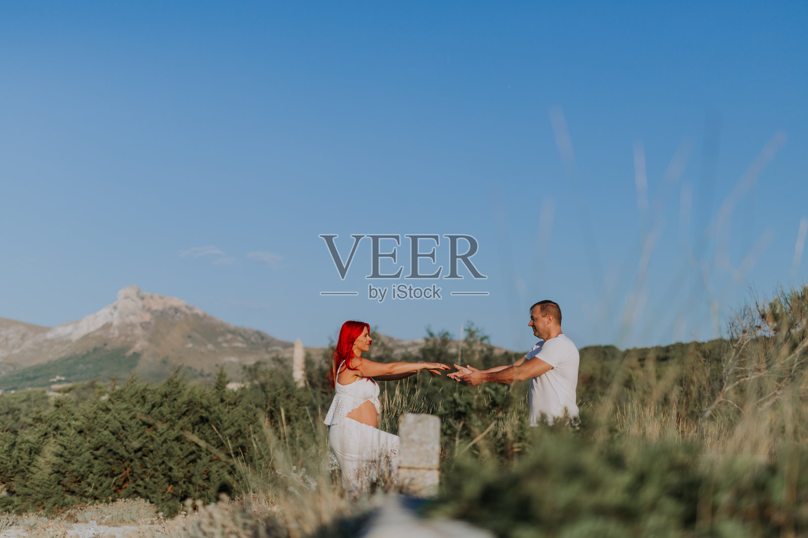 怀孕的情侣在郁郁葱葱的沙丘中走向彼此，伸出手臂温柔地握住对方的手。照片摄影图片