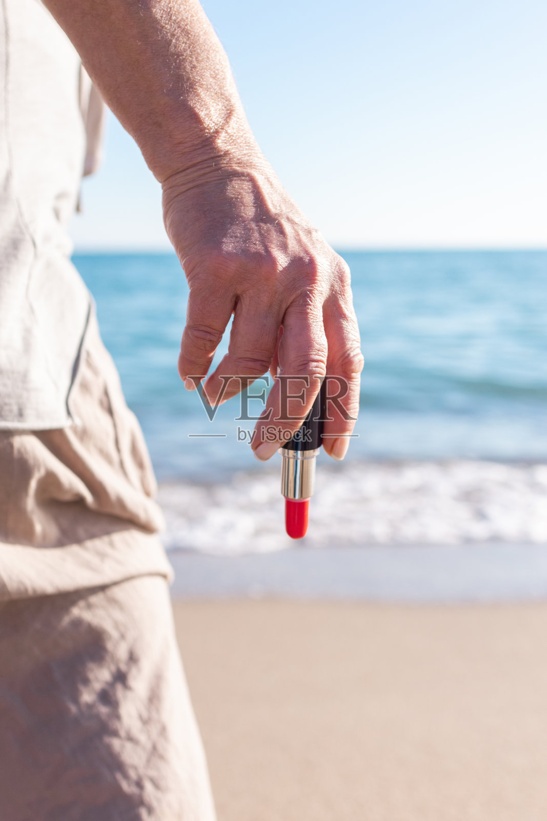 海滩上，一位无法辨认的老年女性背影，手中拿着红色口红，为约会化妆。照片摄影图片