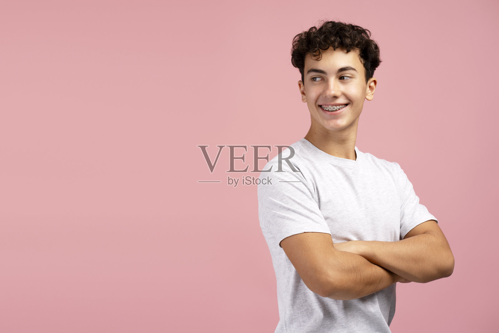 快乐、迷人的青少年男孩，带着牙套，双臂交叉，摆姿势，背景为粉色。照片摄影图片